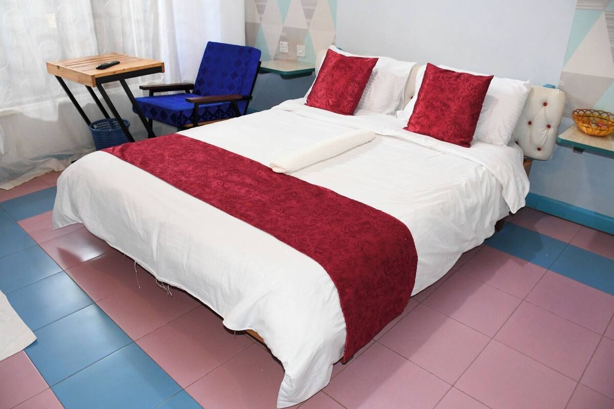 Una cama bien tendida con colcha roja y blanca, una silla azul y una mesita con un control remoto.