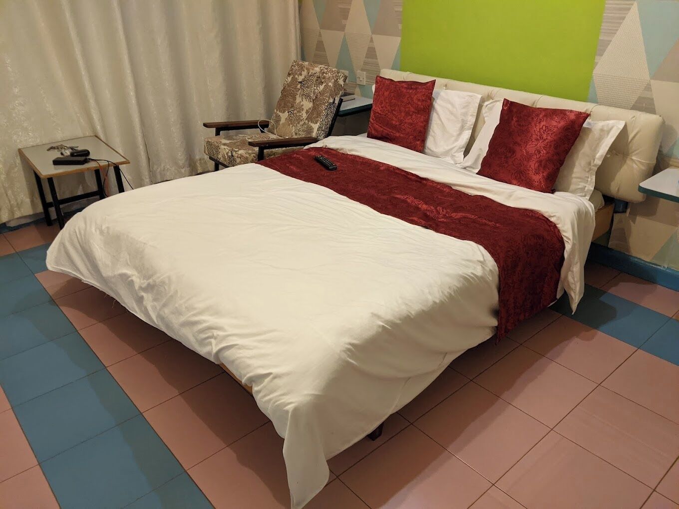 A neatly made bed with a white and red bedspread, a chair with a floral pattern, and a small table with a remote control.