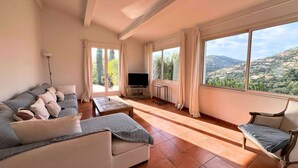Un soggiorno con divano, tavolino, televisione e vista su un paesaggio collinare attraverso ampie finestre.