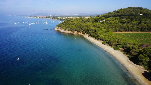 Vista aerea di una zona costiera con acque cristalline, una spiaggia sabbiosa e un promontorio ricoperto da una fitta vegetazione.