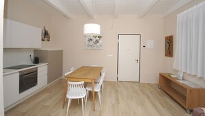 A modern kitchen with white cabinets, a wooden dining table, and white chairs. There is a white door with a seahorse decoration, a white pendant light, and a small wooden shelf with items on it. - MARINAZZURRA  - Goelba (Porto Azzurro)