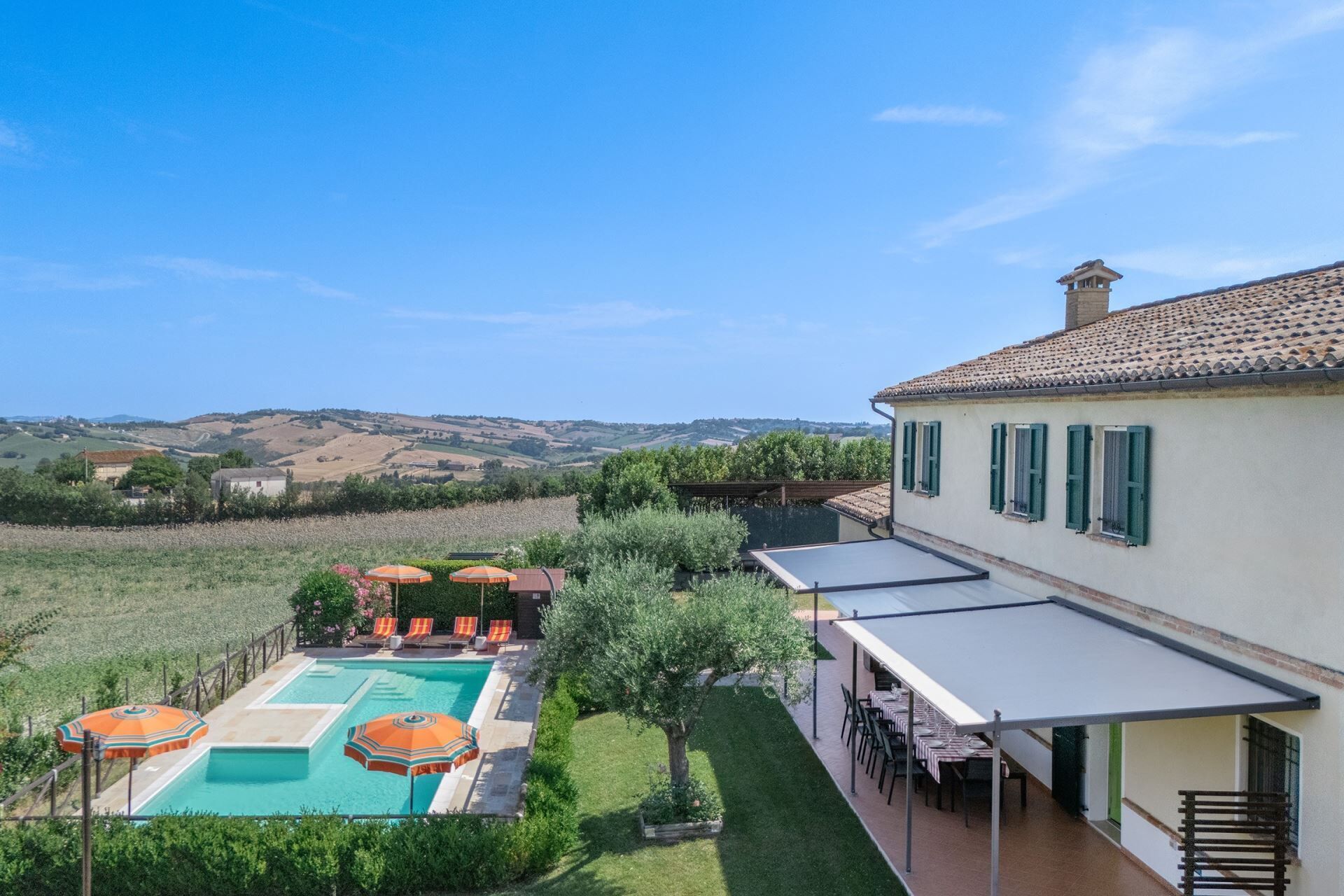 A pool with lounge chairs and umbrellas, a patio with tables and chairs, and a view of rolling hills in the background.