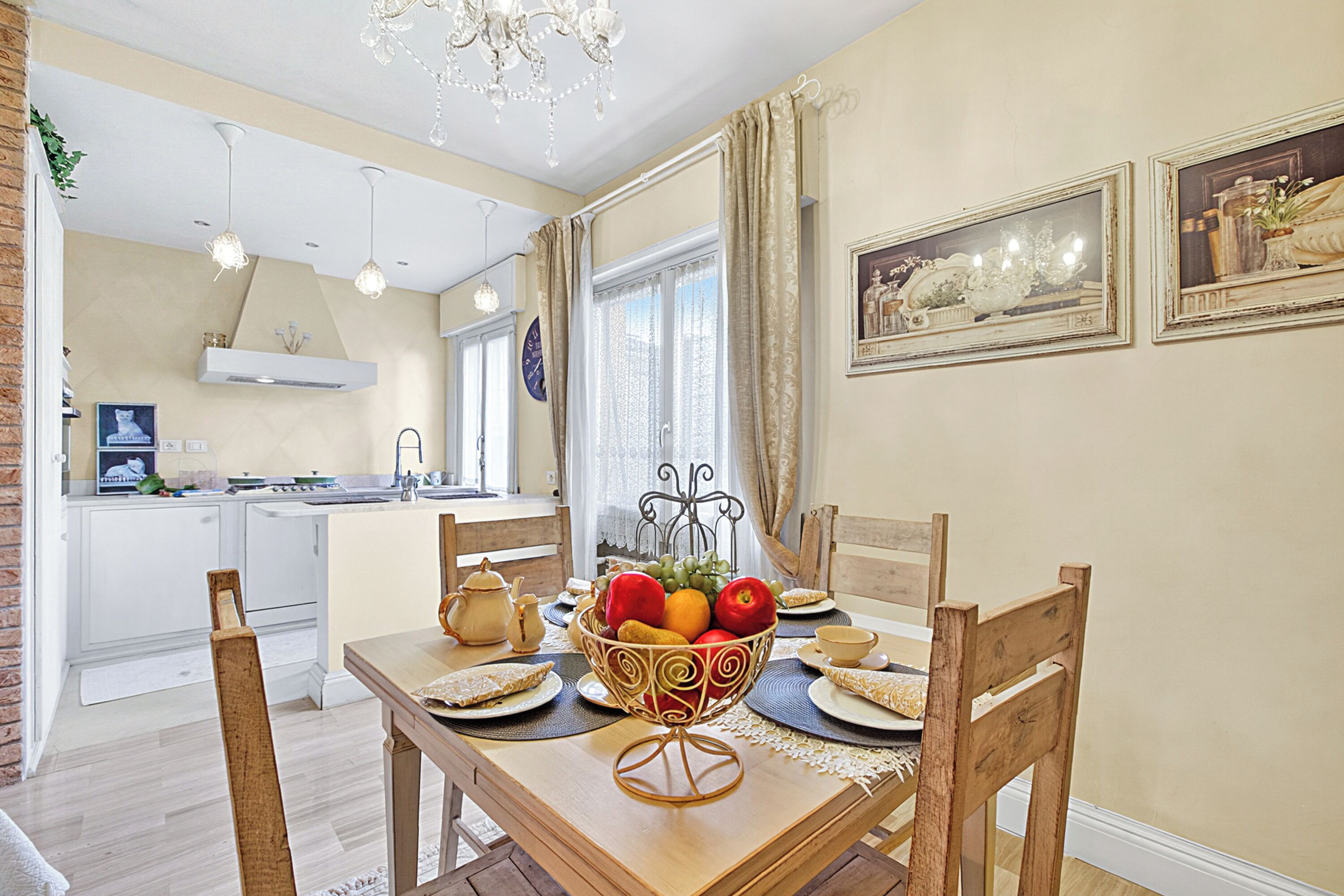 A dining area with a wooden table, chairs, and a fruit bowl. There is a kitchen in the background with white cabinets and a hanging light fixture.