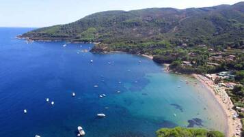 An aerial view of a coastal area with clear blue waters, boats, and a beach with buildings and greenery.