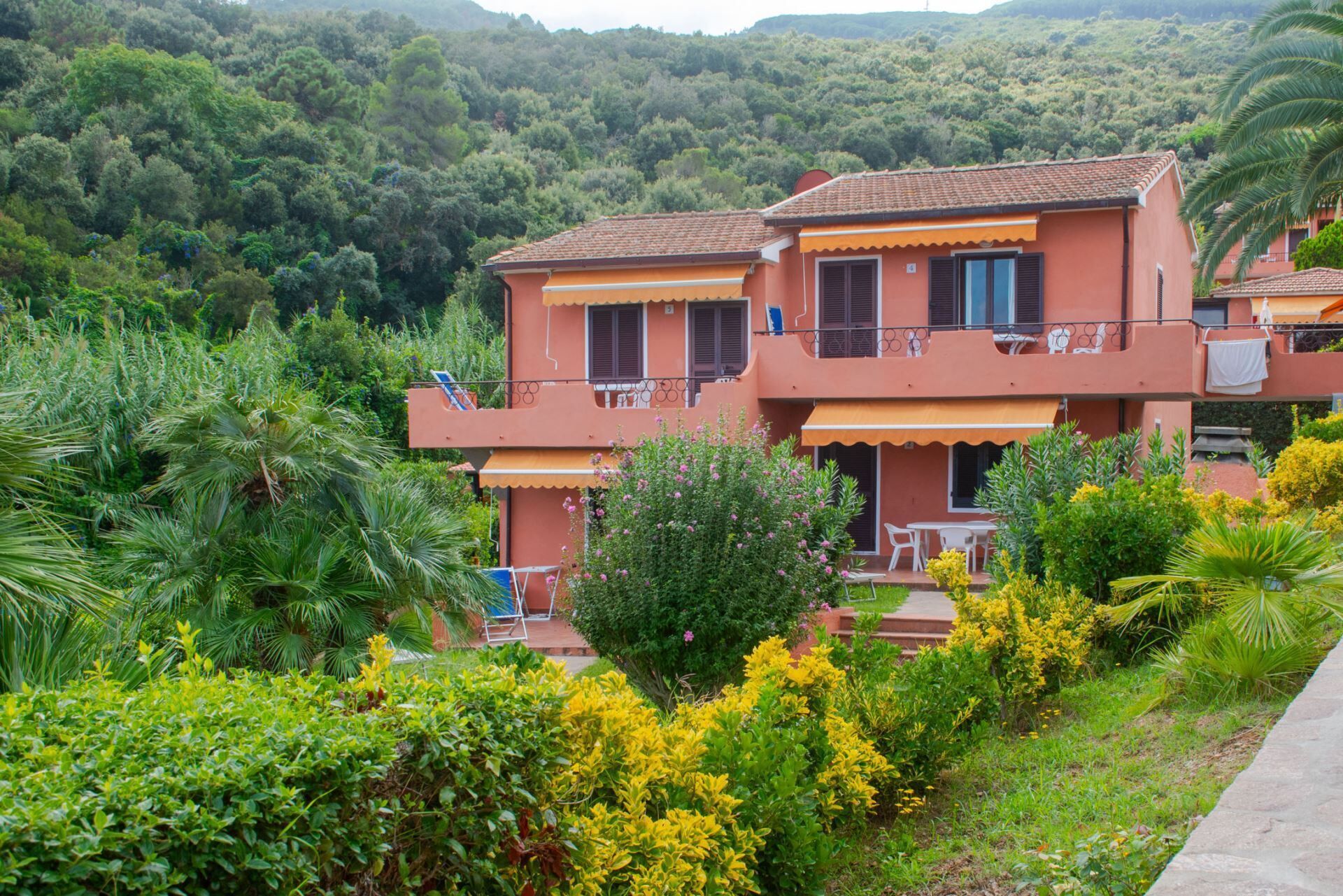 Un bâtiment à deux étages doté d’un balcon, entouré d’une végétation luxuriante et avec des collines en arrière-plan.
