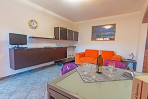 Ein Wohnbereich mit einer Couch, einem Esstisch mit Gläsern und einer Flasche, einem Fernseher auf einem Schrank und einer Uhr an der Wand.