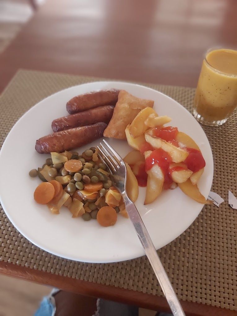 Un plato de comida con salchichas, un alimento frito, verduras mezcladas y un vaso de jugo sobre una mesa.