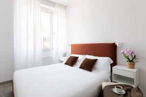 Ein ordentlich bezogenes Bett mit weißen Leinentüchern und zwei braunen Kissen, ein weißer Nachttisch mit einer kleinen Topf-Orchidee und eine Tasse Kaffee auf einem Untersetzer.