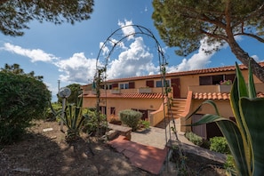 Un edificio de dos pisos con techo de tejas, un camino de piedra que conduce a la entrada y un jardín con diversas plantas y un arco de metal.
