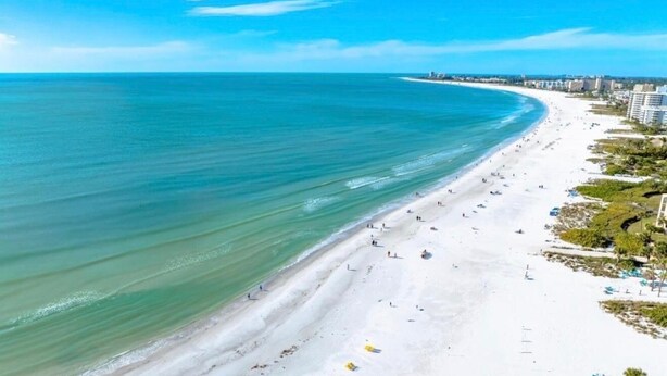 Een strand met een breed zandstrand, helderblauw water en een paar gebouwen op de achtergrond.