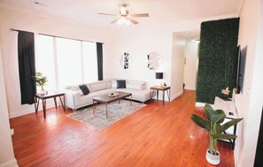 A living room with hardwood flooring, a white sofa, a coffee table, and a ceiling fan. - LUX Home, 3min from LSU, KING Bed (Baton Rouge)