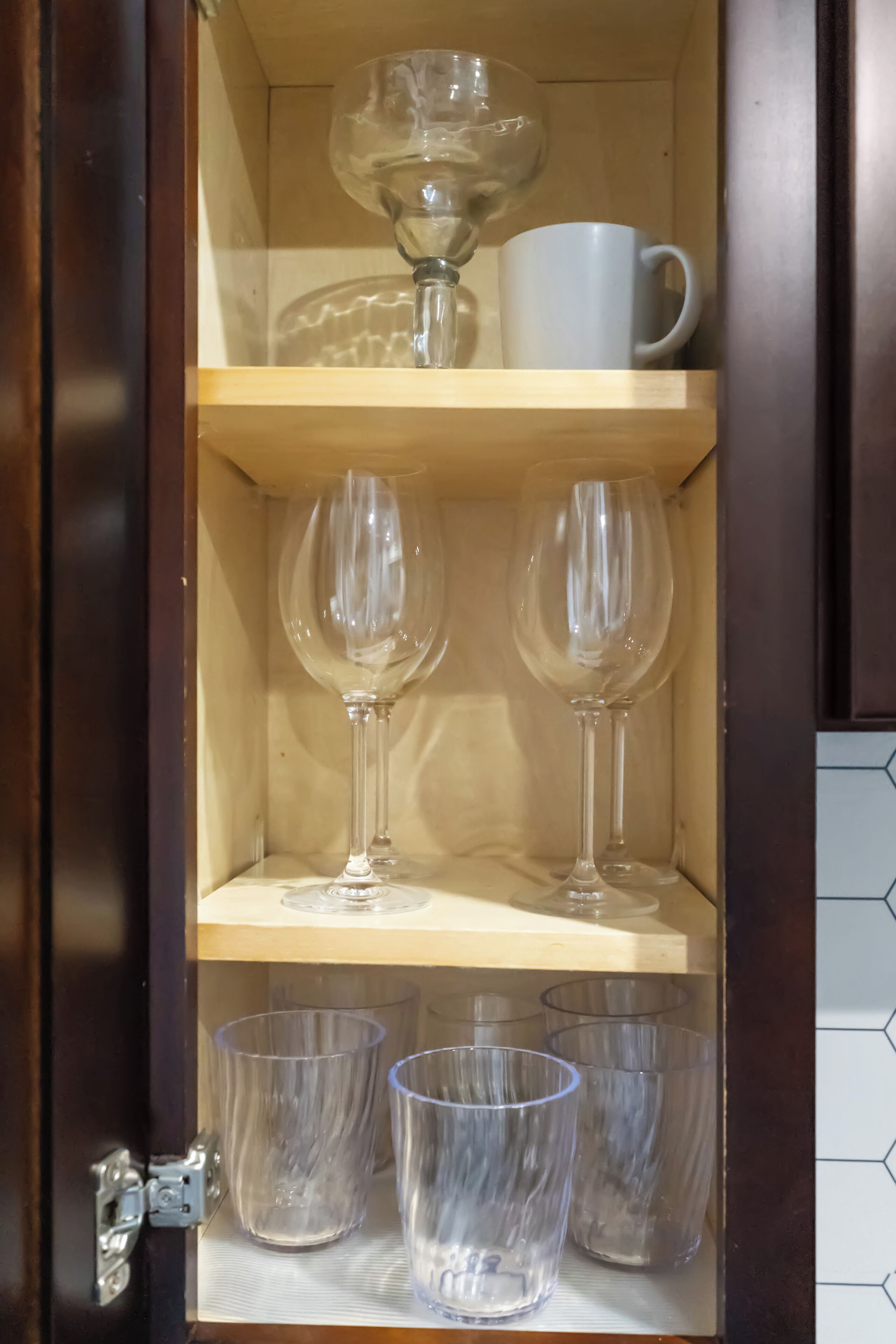 Un meuble en bois avec trois étagères. L’étagère supérieure contient une tasse blanche et deux verres à vin. L’étagère du milieu contient deux verres à vin. L’étagère inférieure contient trois verres transparents.