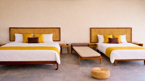 Une chambre d’hôtel avec deux lits, chacun doté d’une tête de lit en osier, de draps blancs et d’éléments décoratifs jaunes. On y trouve des tables de chevet, un banc et un panier en osier posé sur le sol.