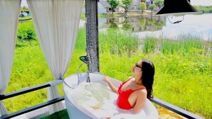 Eine Frau im roten Badeanzug entspannt sich in einer Badewanne mit Blick auf eine Wiese und ein Gewässer.