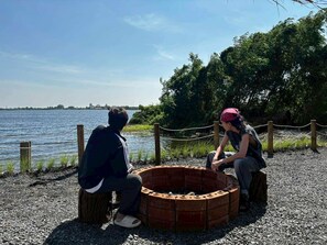 Two people sitting by a circular brick fire pit on a gravel path with a body of water and trees in the background.