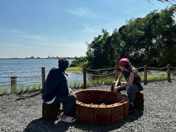 Two people sitting by a circular brick fire pit on a gravel path with a body of water and trees in the background.