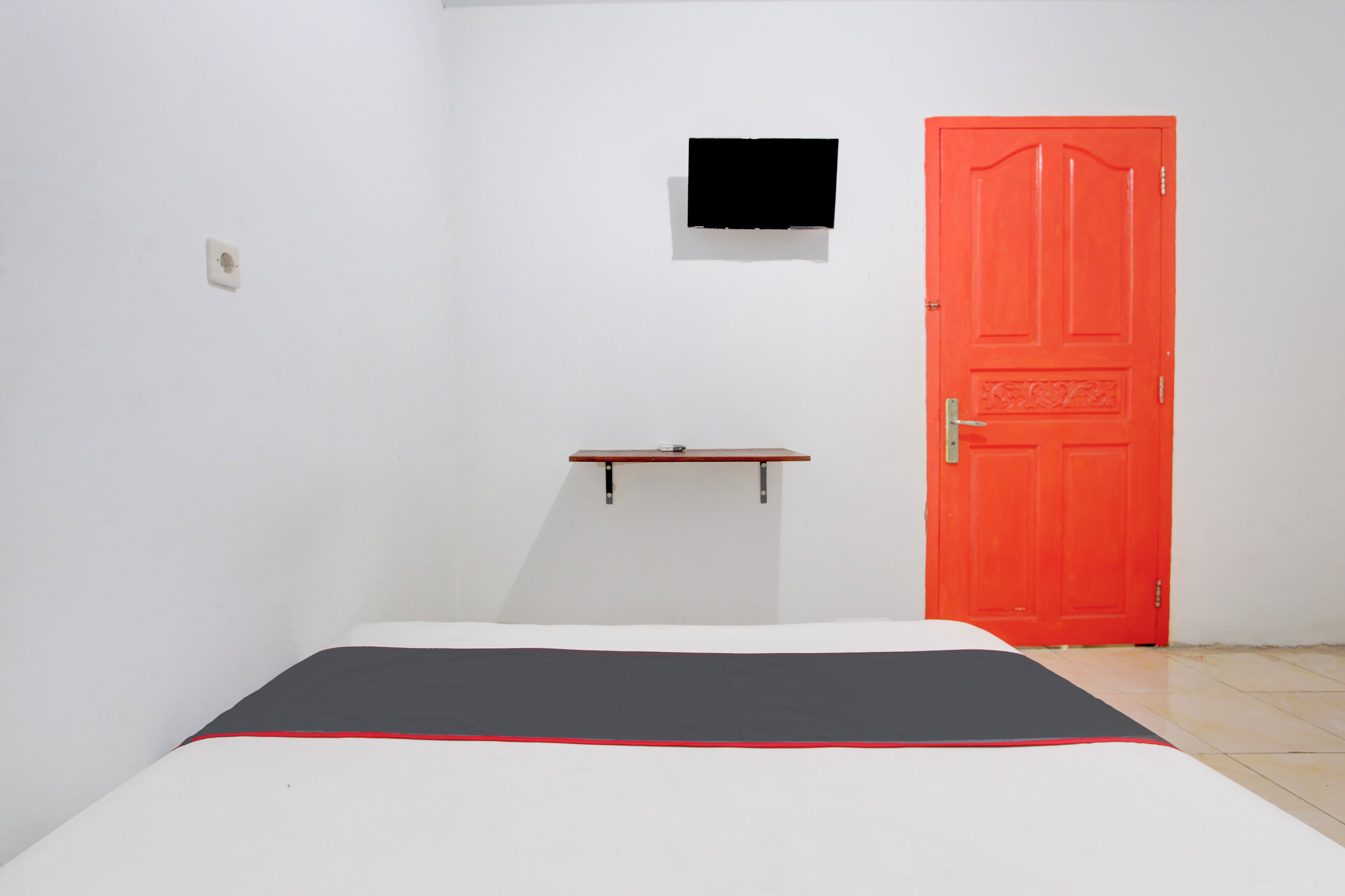 A room with a bed, a red door, a small shelf, and a TV mounted on the wall.
