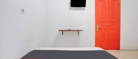 A room with a bed, a red door, a small shelf, and a TV mounted on the wall.