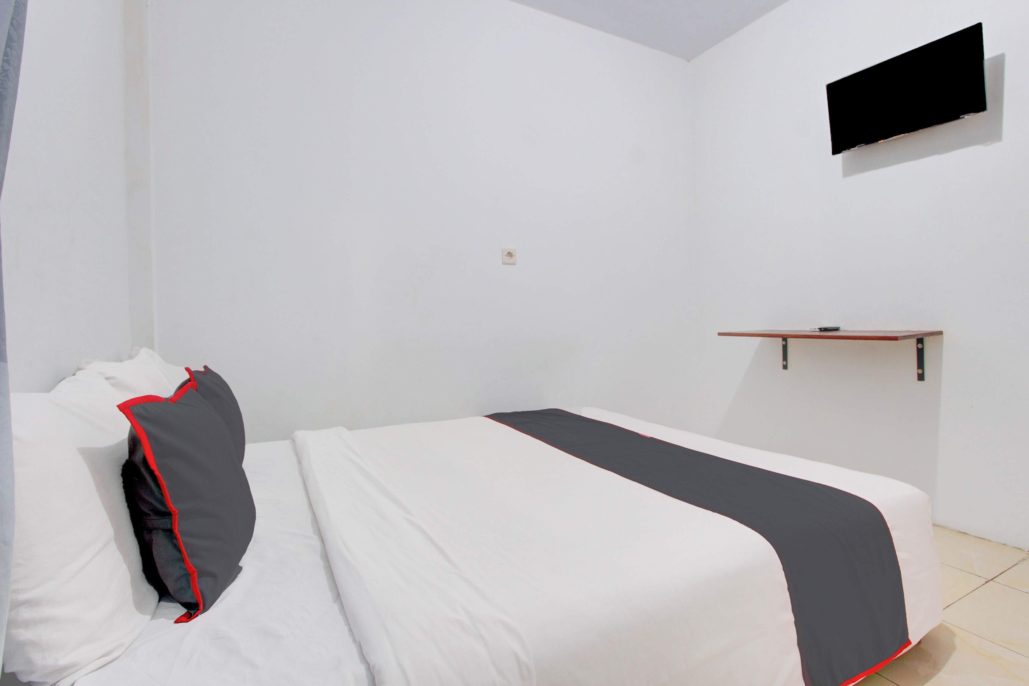 A hotel room with a bed, a flat-screen TV mounted on the wall, and a small wooden shelf.