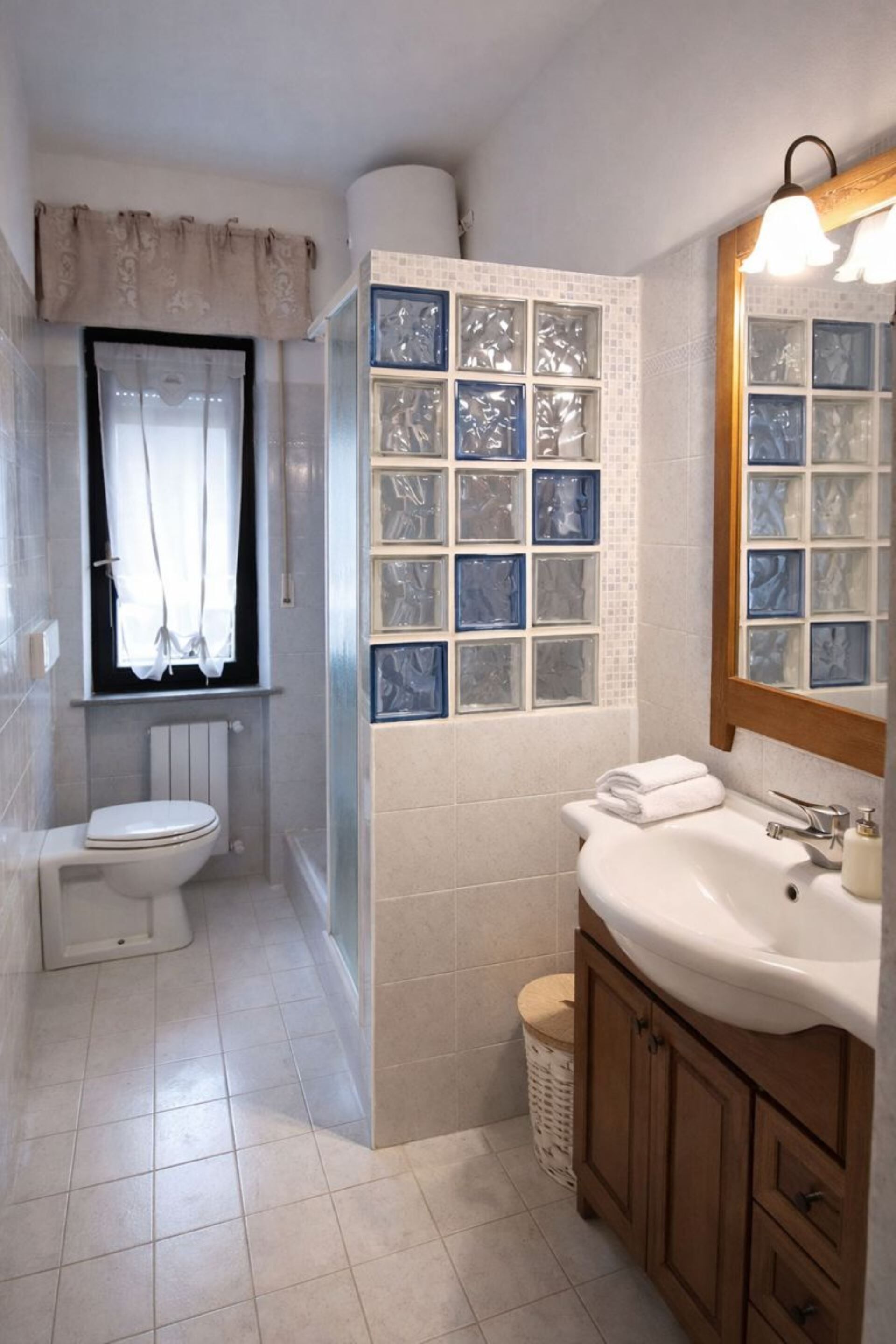 A bathroom with a toilet, sink, and mirror. There is a window with curtains and a tiled wall with decorative glass panels.
