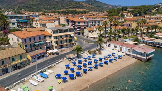 Uma cidade costeira com uma área de praia, apresentando fileiras de guarda-sóis azuis e espreguiçadeiras, edifícios com telhados de terracota e um cenário montanhoso.