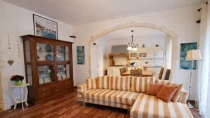 Uma sala de estar com piso de madeira, um sofá listrado, um armário com portas de vidro e uma cozinha ao fundo.
