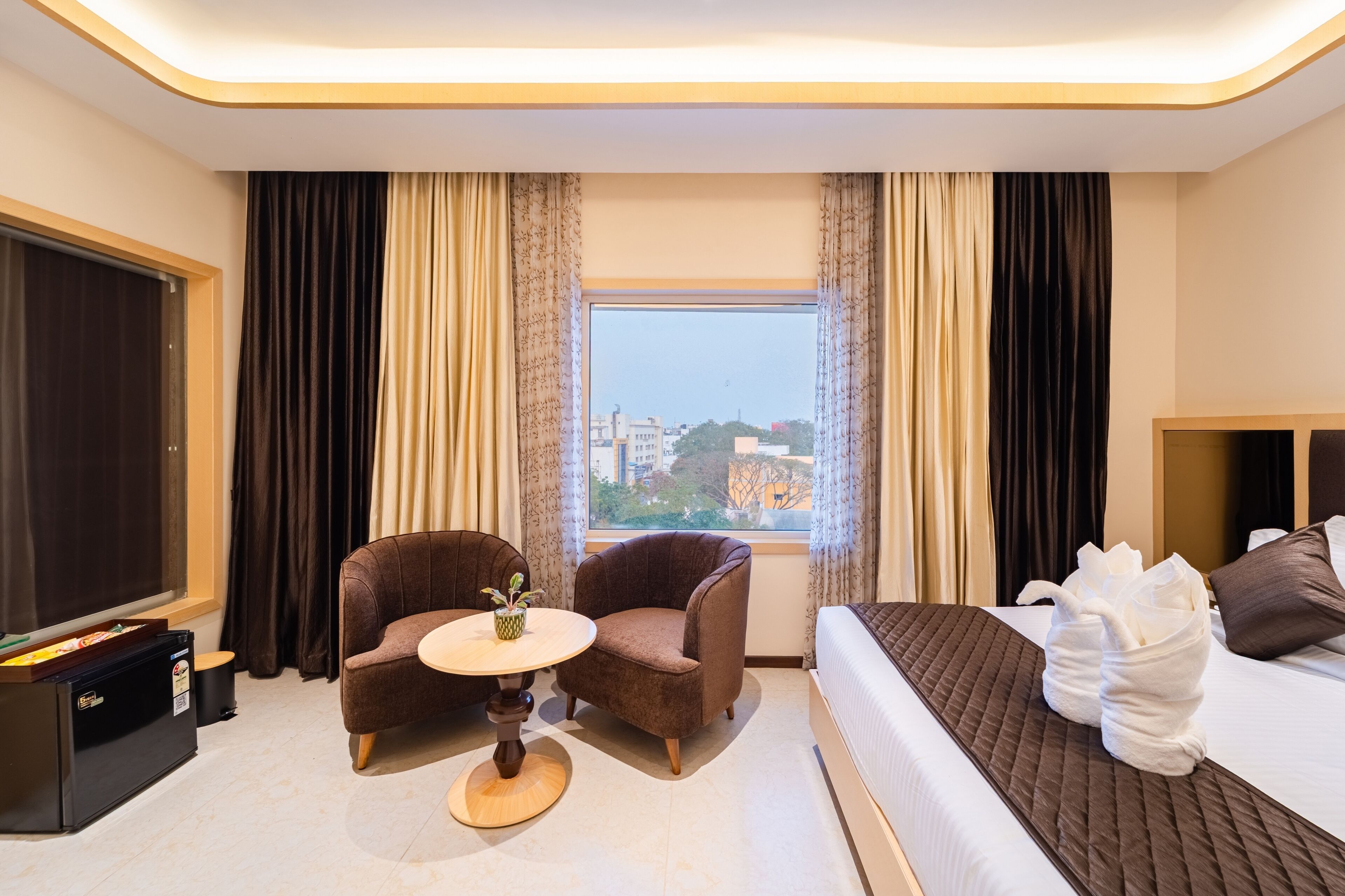 A hotel room with a bed, a small table with a plant, two armchairs, a TV, and a window with curtains.