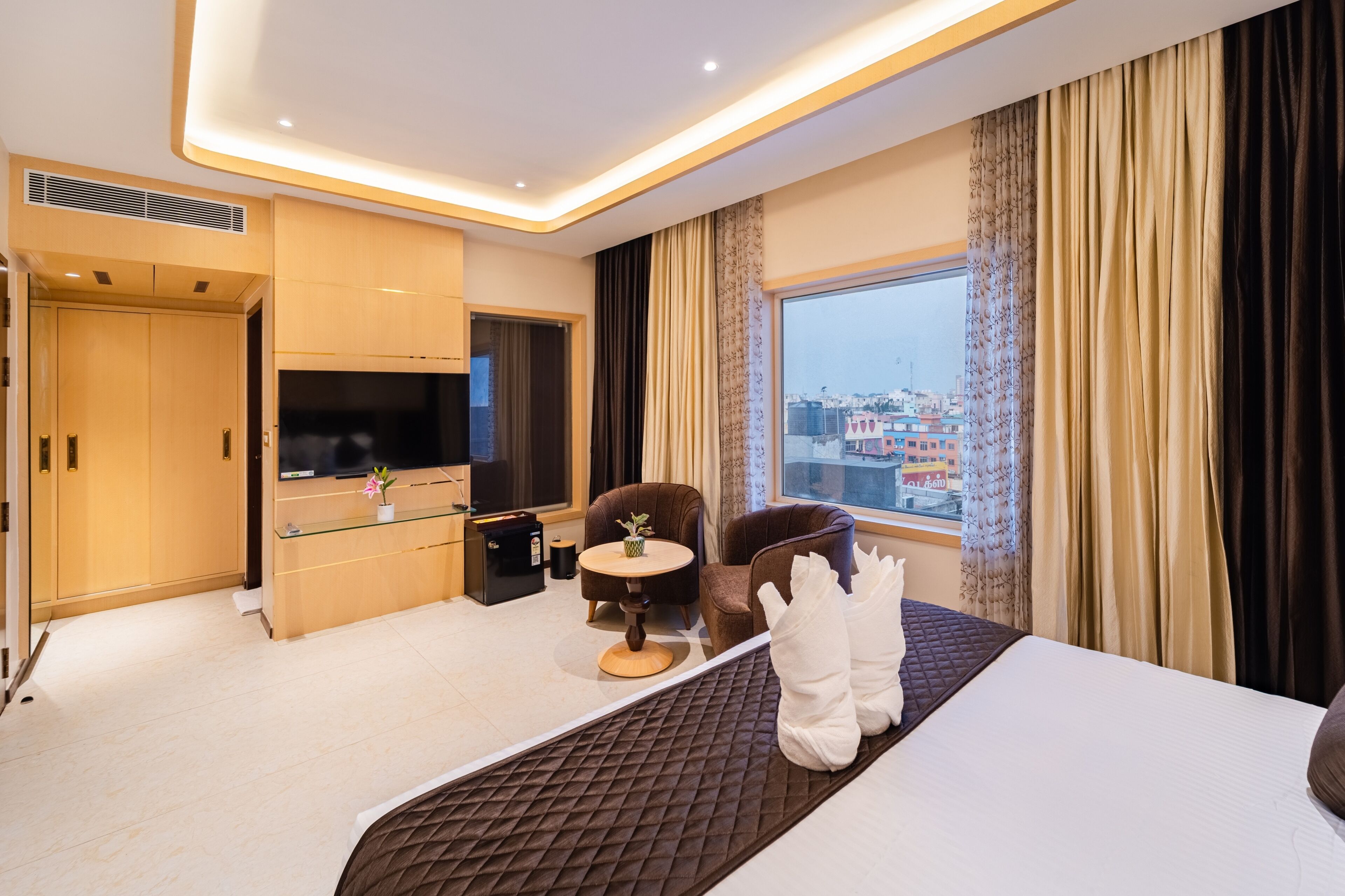 A modern hotel room with a bed, a TV, a seating area with a table, and a view of the city.