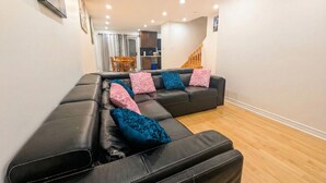 A modern living room with a black leather sectional sofa, wooden flooring, and a staircase leading to an upper level. - Spacious 2BR | FREE Parking + 1.5 Bath | Near YUL (Laval)