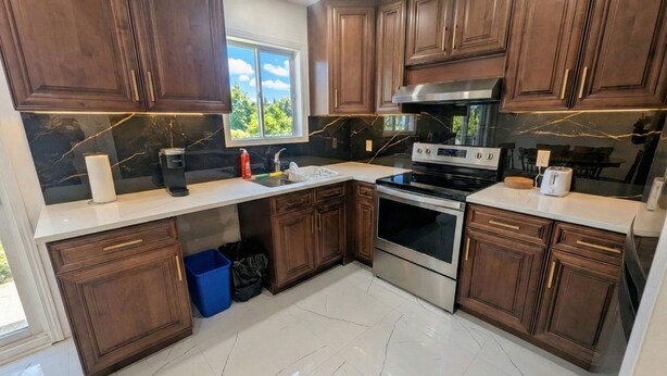 A modern kitchen with dark wooden cabinets, a stainless steel stove, and a large window providing natural light. - Spacious 2BR | FREE Parking + 1.5 Bath | Near YUL (Laval)