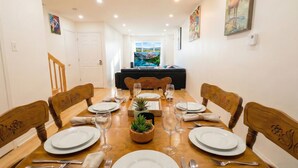 A dining room with a wooden table set for four, surrounded by chairs, with a television and a sofa in the background. - Spacious 2BR | FREE Parking + 1.5 Bath | Near YUL (Laval)