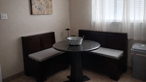 A small room with a round table, two benches, a trash bin, and a window with curtains. - Payne Place 1 (Las Cruces)