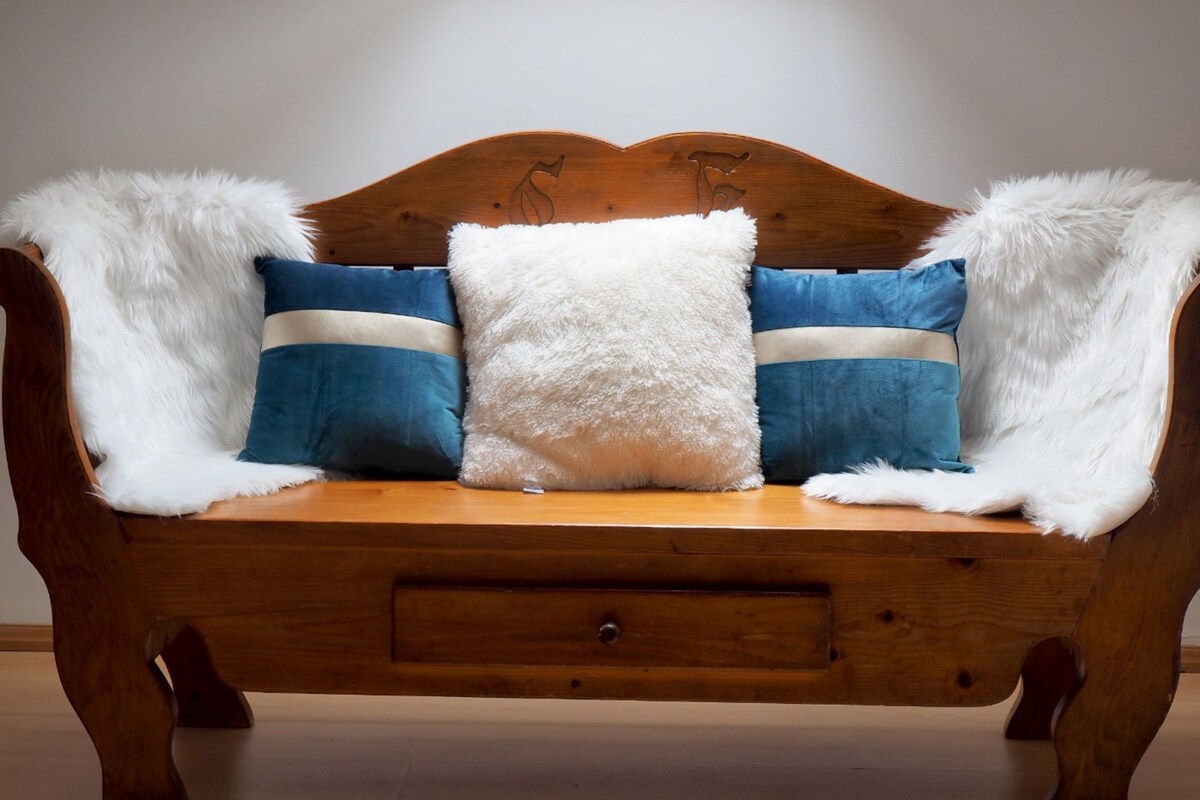 A wooden bench with a white fluffy throw blanket and two blue pillows with white stripes.
