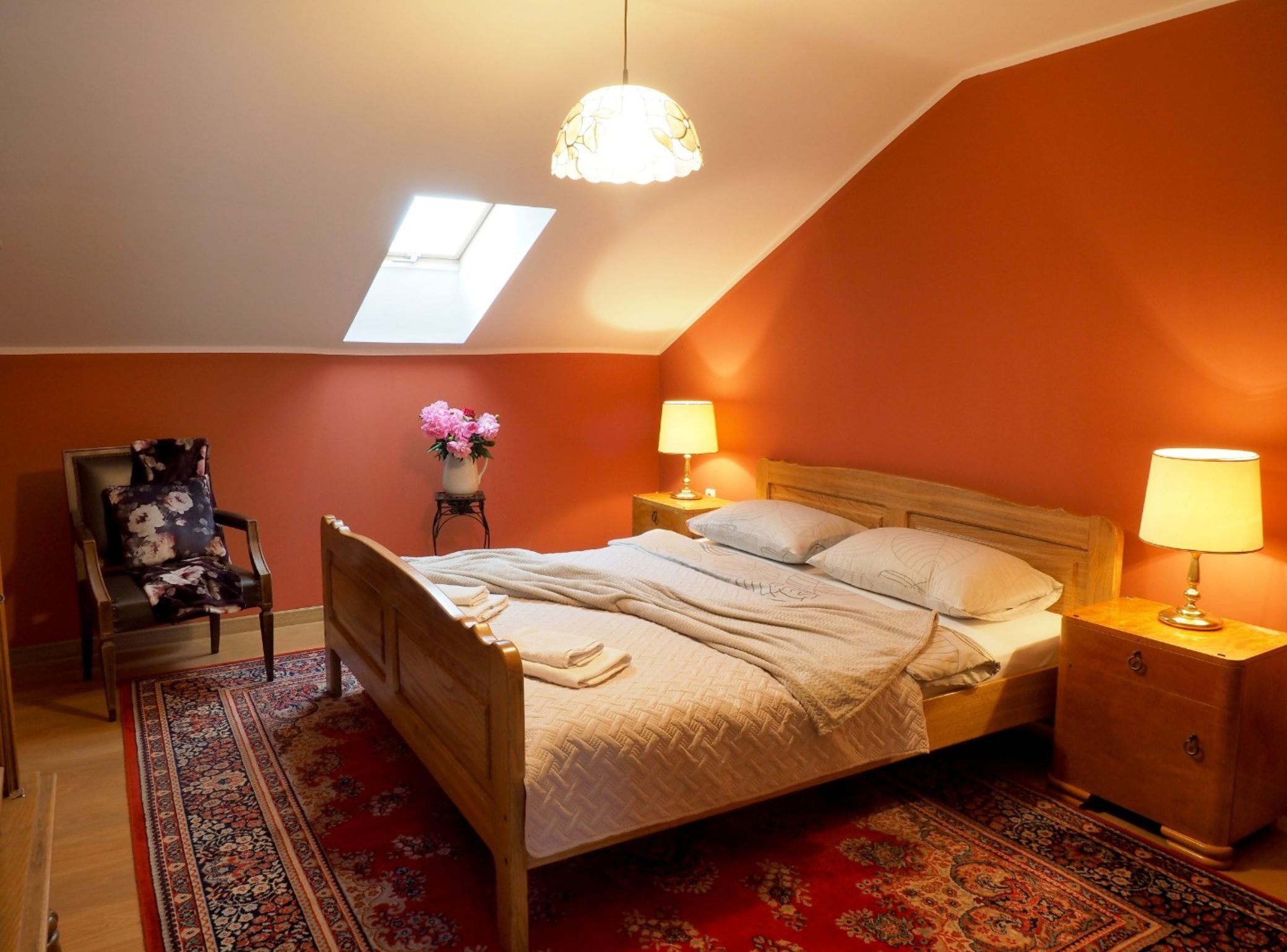 A bedroom with a sloped ceiling, a skylight, a bed with a wooden headboard, two bedside tables with lamps, a chair with a floral cushion, and a red patterned rug.