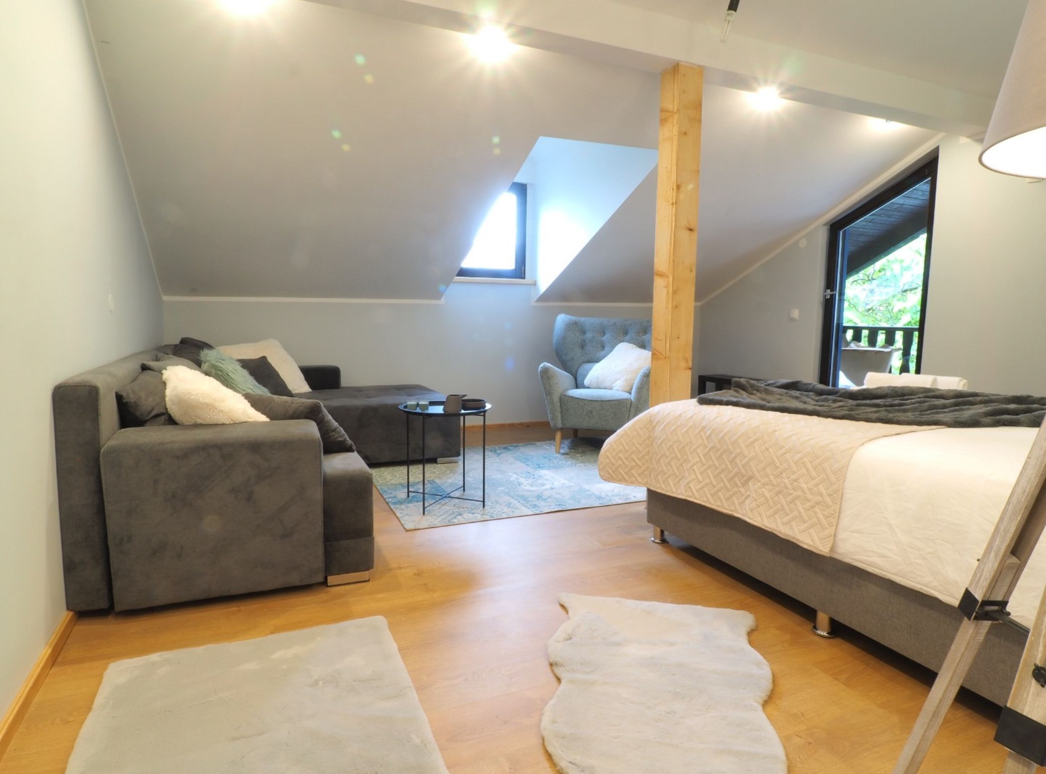 Ett modernt sovrum i loftstil med en grå soffa, en säng med ett vitt täcke, ett litet runt bord och en stol.
