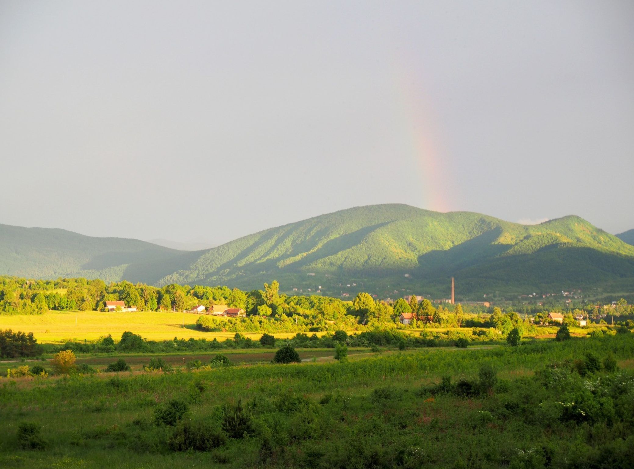 A rural landscape with a rainbow over a mountain, houses in the valley, and fields.
