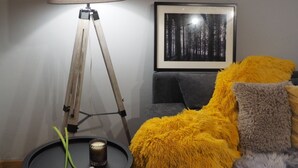 A modern living room with a floor lamp, a framed forest photograph, a sofa with a yellow throw blanket, and a small table with a candle and a white flower. - Apartments & Rooms Latin (Plaski)