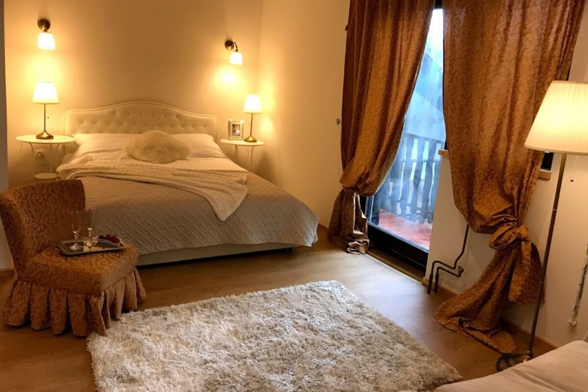 A bedroom with a large bed, two bedside lamps, a chair with a tray, and a view of the outdoors through a window with curtains.
