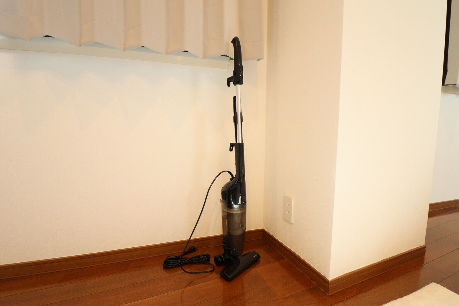 Un aspirateur dans un coin d’une pièce dont le sol est en bois et dont le mur est blanc.
