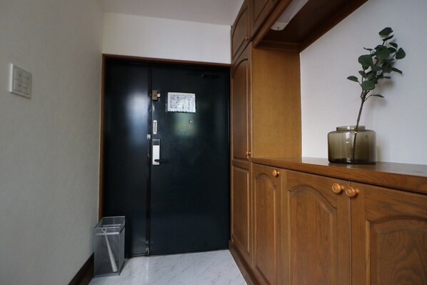 A hallway with a black door, a trash bin, and a wooden cabinet with a plant on top. - Yokohama Mega Stay (Yokohama)