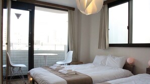 A hotel room with two beds, a white chair, a small table, and a balcony with a view of buildings. - Yokohama Mega Stay (Yokohama)