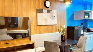 A modern living room with a sofa, dining table, and chairs, a clock on the wall, and a kitchen area with a microwave.