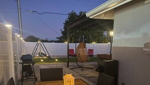 Una veranda esterna con tavolo in legno e sedie in vimini, una poltrona sospesa e un lanterna.