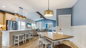 A modern kitchen with a central island, white bar stools, and a wooden dining table. - Beautiful Home | Short Walk To The Beach | Pool (Oak Island)