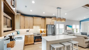 A modern kitchen with wooden cabinets, a central island with stools, stainless steel appliances, and a large window providing natural light. - Beautiful Home | Short Walk To The Beach | Pool (Oak Island)