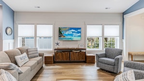 A living room with a beige sofa, a grey armchair, and a wooden cabinet. There is a painting of a beach scene on the wall. - Beautiful Home | Short Walk To The Beach | Pool (Oak Island)