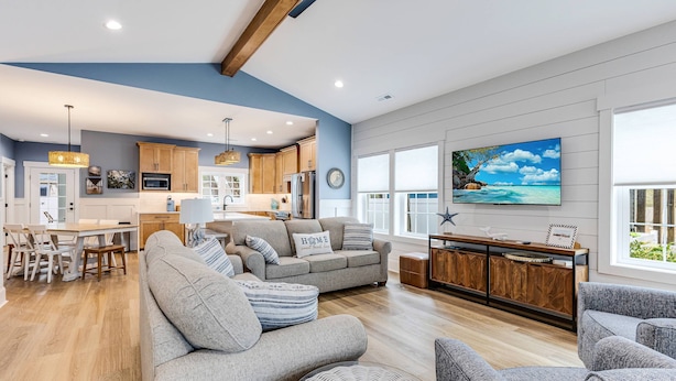 A modern living room with a grey sofa, a dining area with wooden chairs, and a kitchen with wooden cabinets and a large window. - Beautiful Home | Short Walk To The Beach | Pool (Oak Island)