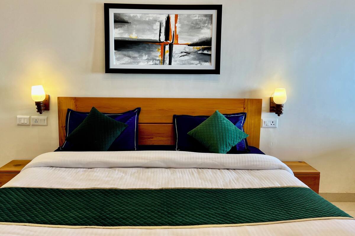 A neatly made bed with a green quilt, two wooden nightstands, a framed abstract painting above the bed, and wall-mounted lamps on either side.