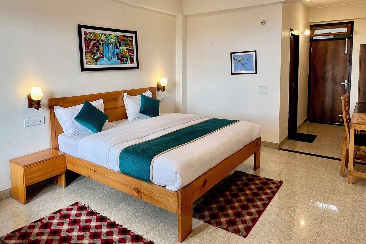 A hotel room with a wooden bed, a small table, a chair, a painting on the wall, and a door leading to another room.