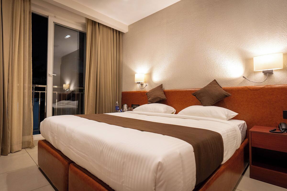 A hotel room with a large bed, a nightstand, a lamp, and a view of the outside through a sliding door.